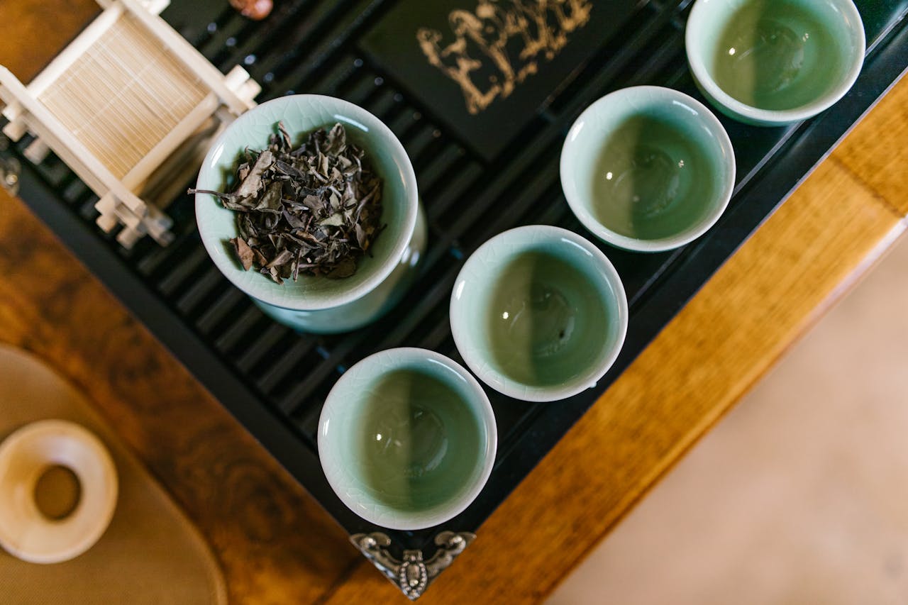 why-choose-us Top view of a traditional tea set with green teacups and dried tea leaves on a bamboo rack, captured in a warm setting.